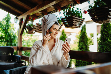 One young caucasian woman is relaxing and taking care of her skin on her balcony with towel on her head and bathrobe while using face roller while using mobile phone
