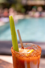 Close up shot of a Bloody Mary cocktail with a celery stick inside and crushed spices on the glass' edge at the pool, in a sunny afternoon in Ibiza. Vacation mood, relax, pool party vibes.