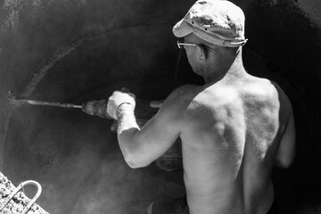 a young muscular tanned male worker with a hammer drill in his hands punches a hole in a reinforced...