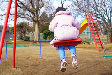 寒い日も元気に外遊び。冬の公園でブランコに乗っている女の子の後ろ姿