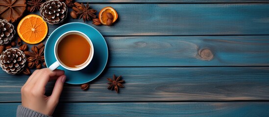 A warm and inviting autumn or winter scene with a cup of coffee cinnamon sticks sugar orange slices and pine cones arranged on a wooden background Blue mittened hands hold the coffee cup creating a c - Powered by Adobe