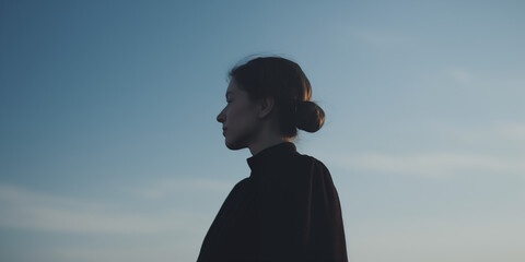 Young woman with dark hair in a bun looks off into the distance against a clear sky, creating a serene and reflective atmosphere
