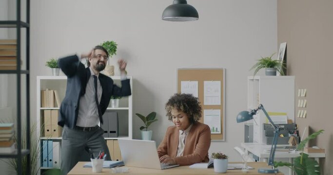 African American businesswoman working with laptop computer while Middle Eastern businessman dancing in office. Workplace and business activity concept.