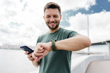 A man smiles while checking his watch and holding a phone, standing by a waterfront with a bridge in the background.