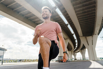 A man performs a knee lift stretch under a bridge, smiling as sunlight filters through
