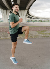 A man warms up under a bridge, lifting his knee high in a dynamic stretching exercise.