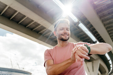 A man checks his watch under a bridge, smiling in the sunlight after a workout session