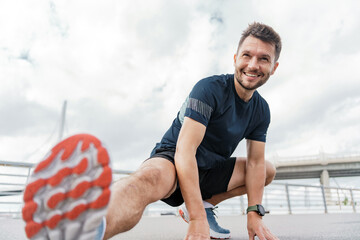 A man stretches his leg while smiling during an outdoor workout, with a bridge in the background.