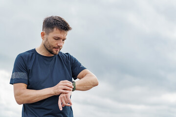 A man checks his watch during an outdoor workout session, with a cloudy sky in the background.