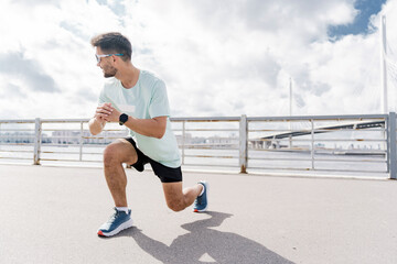 A man in a mint green shirt and black shorts performs a lunge exercise by the waterfront under a cloudy sky.