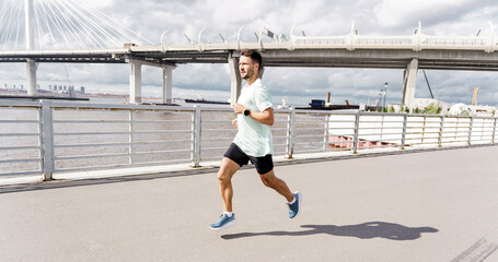 A man in a mint green shirt and black shorts runs by the waterfront under a cloudy sky with a bridge in the background.
