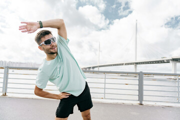A man in a mint green shirt and black shorts performs a side stretch by the waterfront under a cloudy sky.