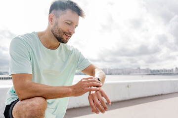 Man sitting on a ledge, checking his smartwatch, wearing a light blue shirt and black shorts