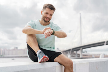 Man checking his smartwatch, wearing a dark shirt and black shorts, against a cloudy sky.