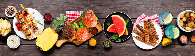 Summer BBQ food table scene. Hamburgers, meat skewers, potatoes, fruit and snacks. Top view on a dark wood banner background.