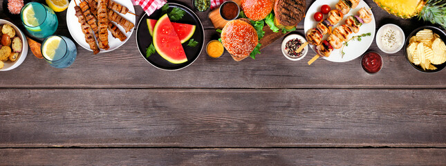 Summer BBQ food top border. Hamburgers, meat skewers, potatoes, fruit and snacks. Overhead view on a dark wood banner background. Copy space.