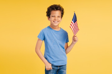 Smiling African American boy, patriot holding American flag looking at camera on yellow background