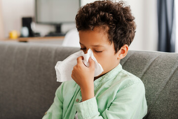 Sick, sad African American boy has runny nose, holding napkin, sitting on comfortable sofa at home
