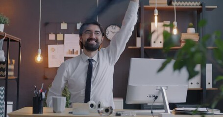 Excited Middle Eastern businessman having fun with jacket and smiling sitting at desk in office at night. Positive emotion and business concept.