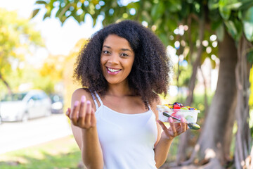 Young African American woman holding a bowl of fruit at outdoors inviting to come with hand. Happy...