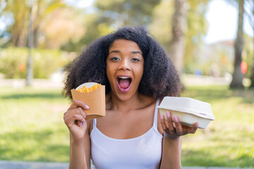 Young African American woman at outdoors taking fried chips and takeaway box food with surprised...
