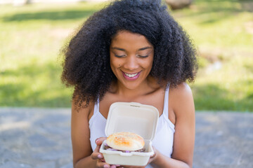 Young African American woman at outdoors holding a burger with happy expression