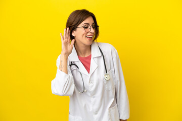 Doctor woman isolated on yellow background listening to something by putting hand on the ear