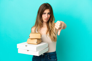 Caucasian woman holding pizzas and burger isolated on blue background showing thumb down with...