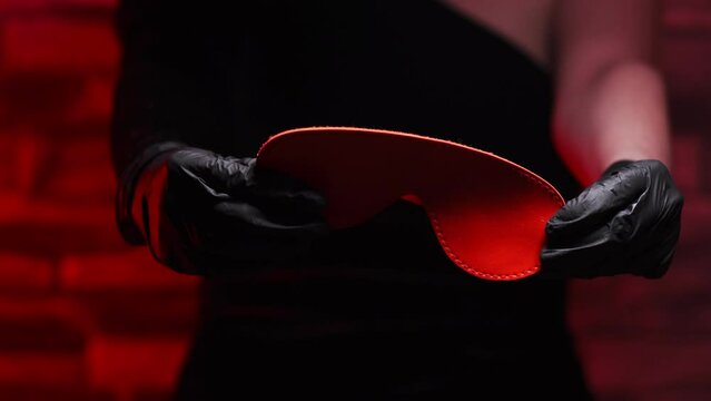 Close-up of female hands in black latex gloves holding a red BDSM headband on a red background. BDSM - outfit for adult sex games
