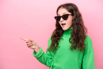 Little caucasian girl wearing sunglasses isolated on pink background surprised and pointing finger...