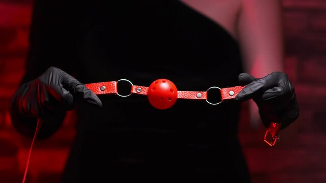 Close-up of female hands in black latex gloves holding a red BDSM gag on a red background. BDSM - outfit for adult sex games
