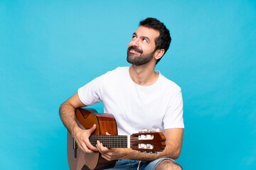 Young man with guitar over isolated blue background looking up while smiling
