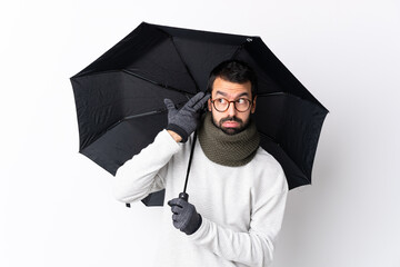 Caucasian handsome man with beard holding an umbrella over isolated white wall with problems making...