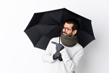 Caucasian handsome man with beard holding an umbrella over isolated white wall pointing to the side...