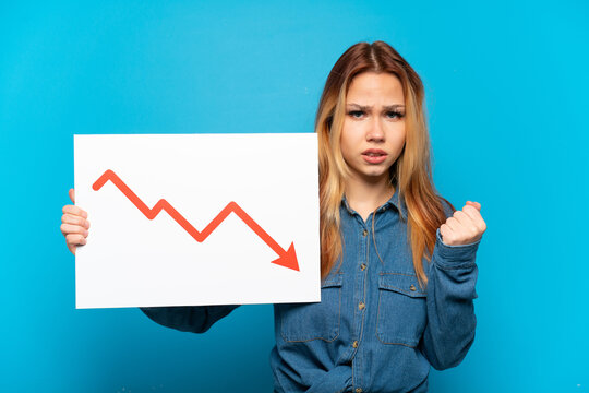 Teenager girl over isolated blue background holding a sign with a decreasing statistics arrow symbol and angry