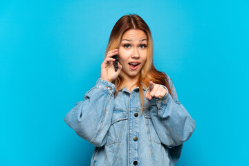 Teenager girl using mobile phone over isolated background surprised and pointing front