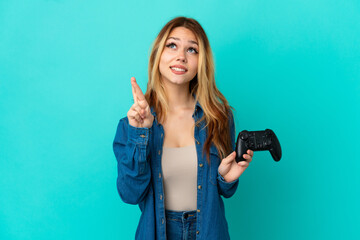 Teenager blonde girl playing with a video game controller over isolated wall with fingers crossing...