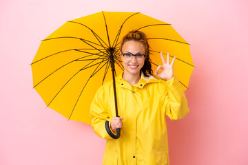 Teenager Russian girl with rainproof coat and umbrella isolated on pink background showing ok sign...