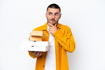 Young caucasian man holding pizzas and burgers isolated on background having doubts and thinking