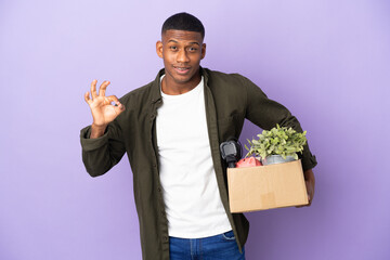 Latin man making a move while picking up a box full of things showing ok sign with fingers