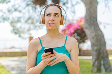Young pretty woman at outdoors listening music with a mobile and thinking