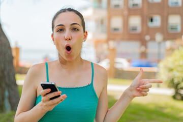 Young pretty woman using mobile phone at outdoors surprised and pointing side