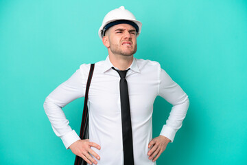 Young architect caucasian man with helmet and holding blueprints isolated on blue background suffering from backache for having made an effort