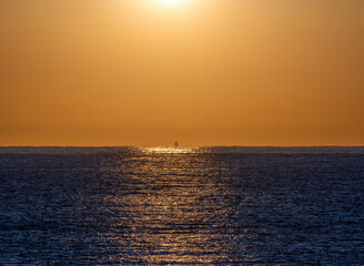 First lights of dawn backlit and illuminating the silhouette of a fishing boat sailing through the calm blue waters of the Mediterranean Sea with the brightness of the sun reflected in its waters.