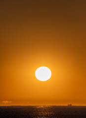 Orange sky with a bright yellow dawn sun illuminating a fishing boat sailing through the Mediterranean Sea and the first rays of the dawn sun creating optical illusions and mirages on the horizon.