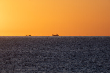 Silhouettes of two fishing boats illuminated by the dawn sun on the horizon of the Mediterranean Sea going out to fish for shrimp, under an orange sky.