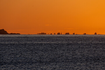 Fishing boats on the horizon line of the Mediterranean Sea heading out to sea from the port and coast of Palamós for shrimp fishing, illuminated by the dawn sun creating mirages and optical illusions.