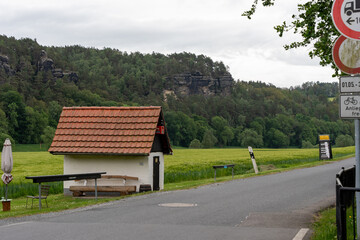 A small red house with a white roof sits in a field. The house is surrounded by trees and grass