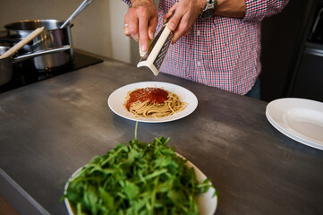 Close-up hands holding grater, grating cheese while seasoning the freshly boiled spaghetti, preparing healthy meal according to Italian recipe. Young man cooking pasta for dinner in the home kitchen.