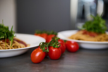 Focus on yummy bunch of ripe organic cherry tomatoes, near two white plates with Italian spaghetti pasta garnished with green leaves of arugula, on the kitchen table. Food background. Italian cuisine